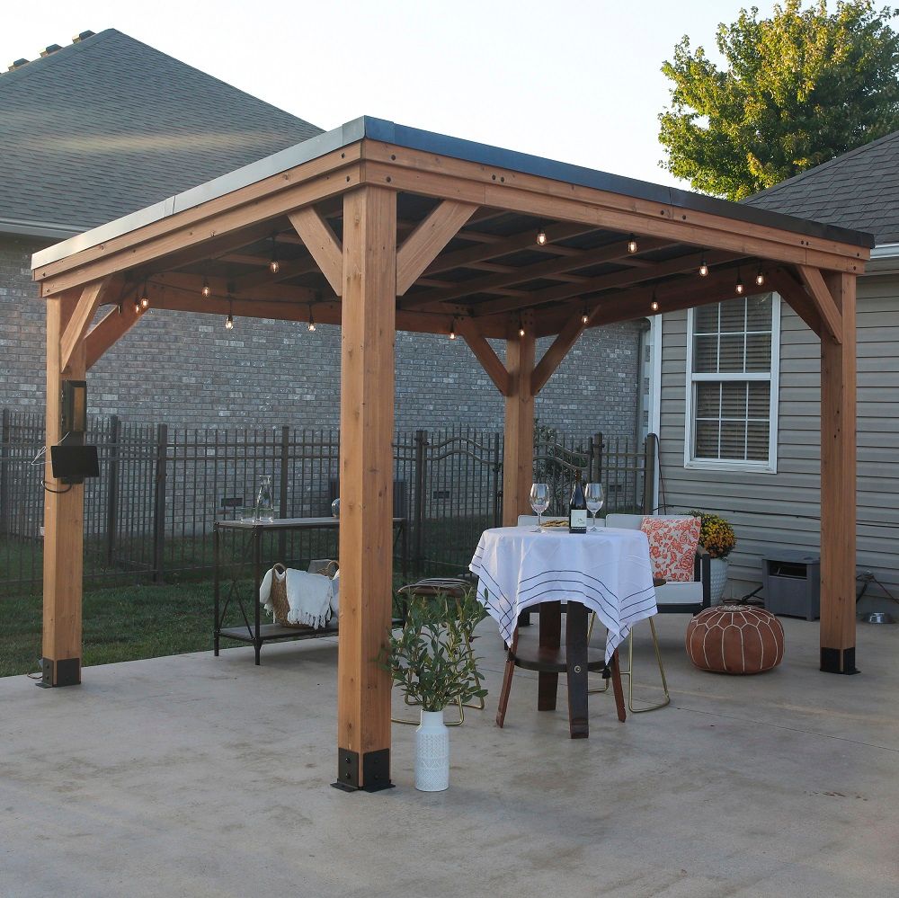 Wooden pavilion on a patio with string lights, a small table, and autumn decor beside a brick house.