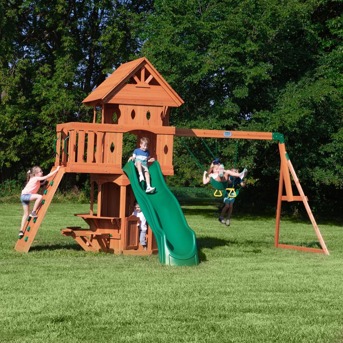 Children playing on a wooden backyard playground with a green slide and swings in a grassy yard.