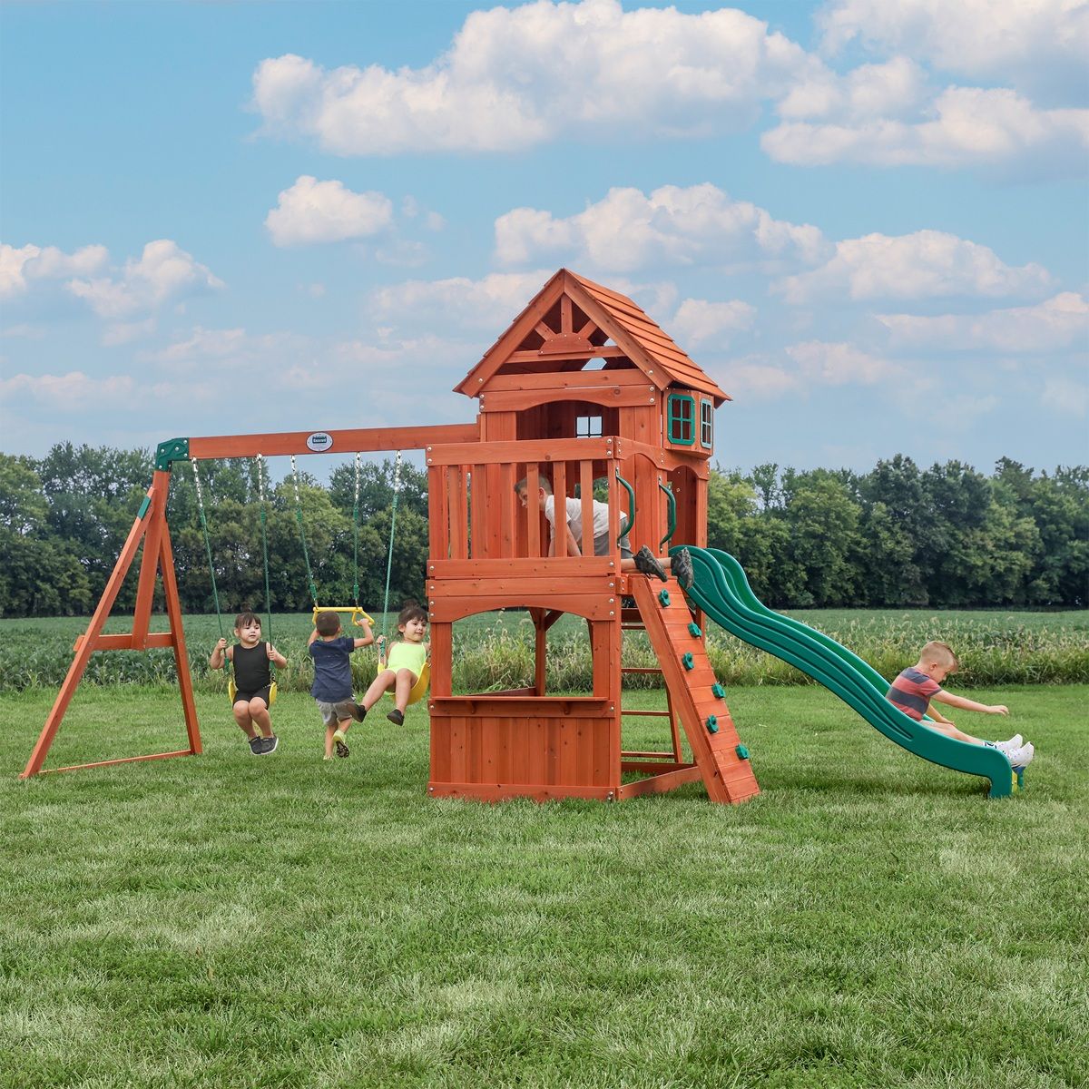 Children playing on a wooden backyard playset with swings and a green slide in a grassy field under a blue sky