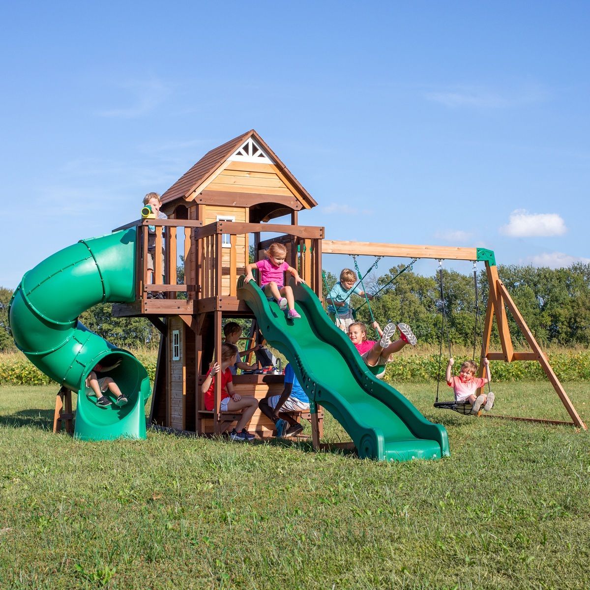 Children playing on a wooden backyard playset with green slides and a swing under a blue sky