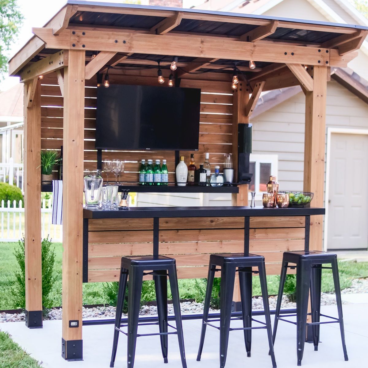 Wooden outdoor bar with black stools, shelves of bottles, and a TV under a roofed patio.