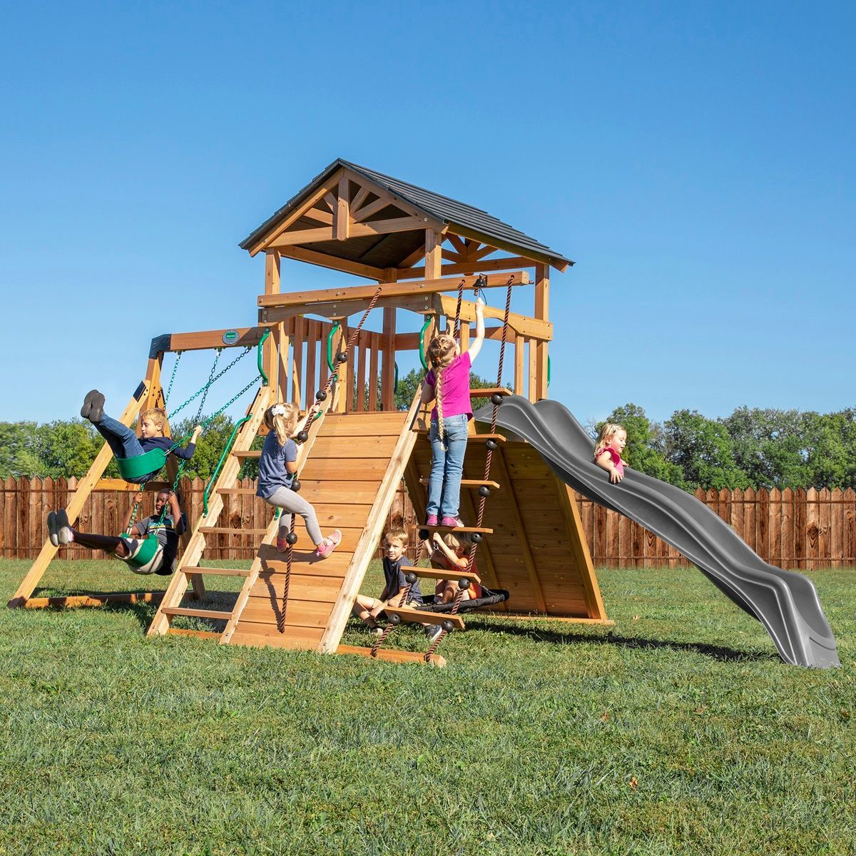 Children playing on a wooden playground with swings, slides, and climbing stairs in a grassy field