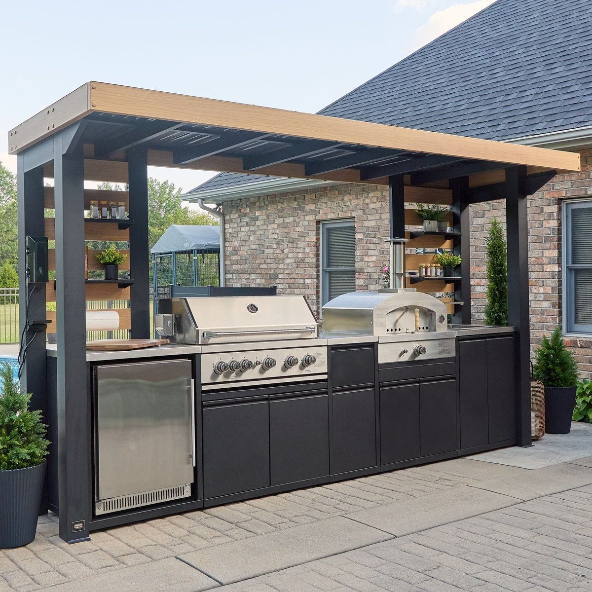 Covered outdoor kitchen with grill, sink, and storage cabinets under a dark pergola on a patio.