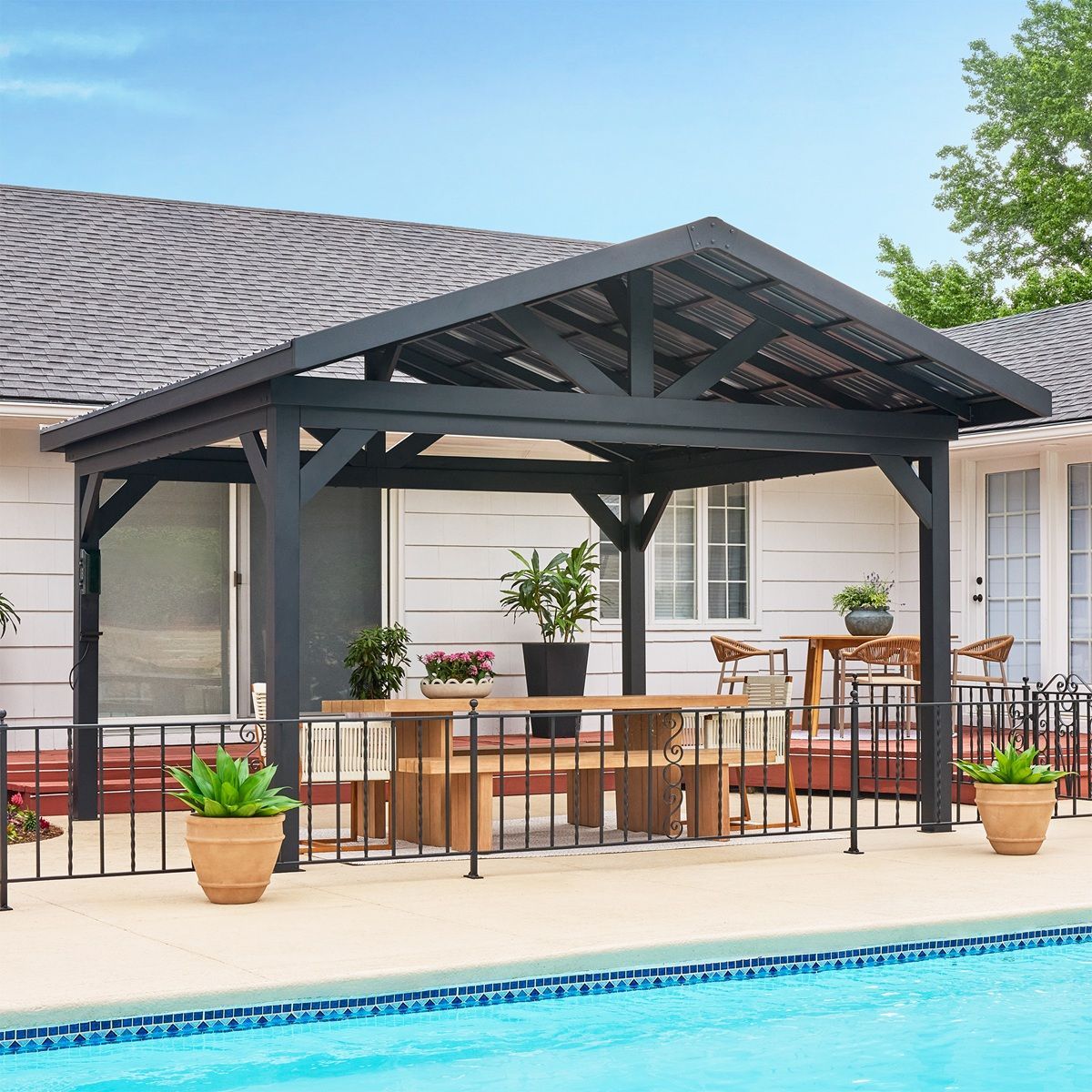 Backyard patio with covered outdoor dining area, black pergola, potted plants, and swimming pool beside a house