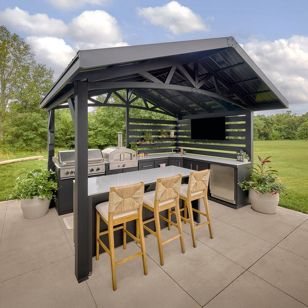 Covered outdoor kitchen with bar stools, grill, and TV on a patio overlooking a grassy field
