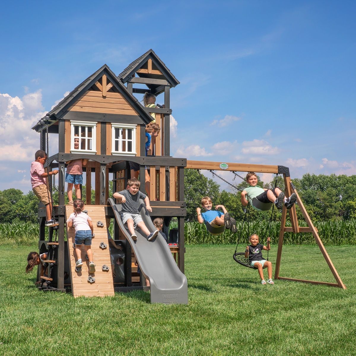 Children playing on a wooden backyard playset with slide, swings, and climbing wall on a grassy lawn.