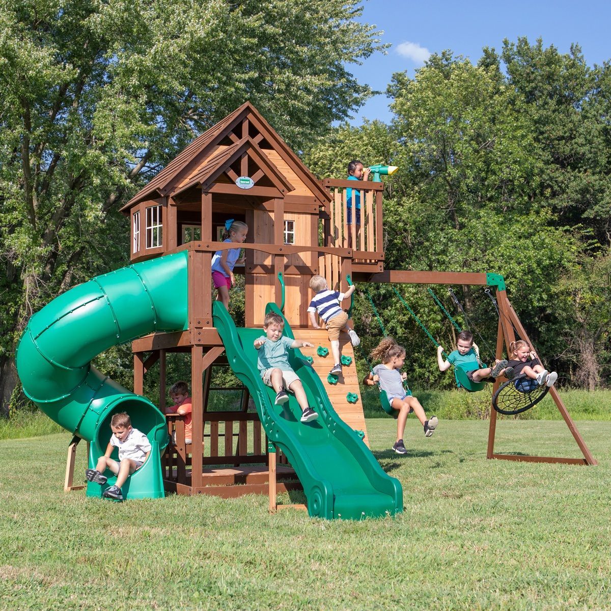 Children playing on a wooden backyard playset with green slides and swings in a grassy yard.