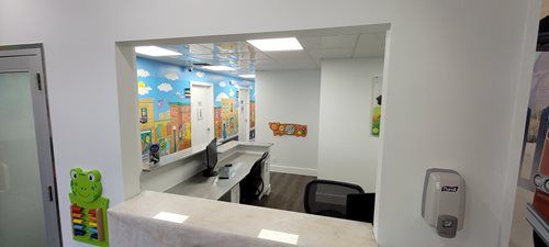 Interior view of a pediatric clinic's reception area. A window-like opening reveals a counter, computer, and colorful murals.