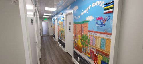 Hallway with Sesame Street mural.  Elmo, a blue monster, and buildings are depicted. Doors line the sides.