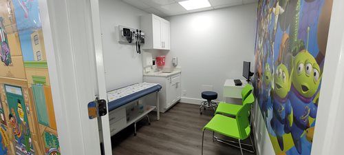 Doctor's office exam room with colorful cartoon wall art, examination table, and two green chairs.