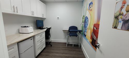 A medical examination room with white cabinets, a computer, and a blood draw chair; a colorful mural is on the wall.