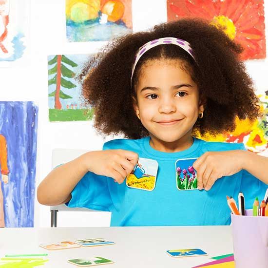 Young girl holding two small, square paintings, smiling, with art in background.
