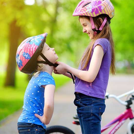 Two children in helmets talk beside a bicycle on a sunny path