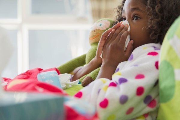 Child in bathrobe blows nose with tissue; stuffed animal and box of tissues nearby.