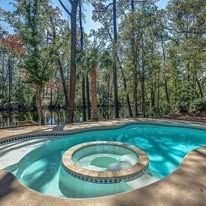 A large swimming pool with a hot tub in the middle surrounded by trees.
