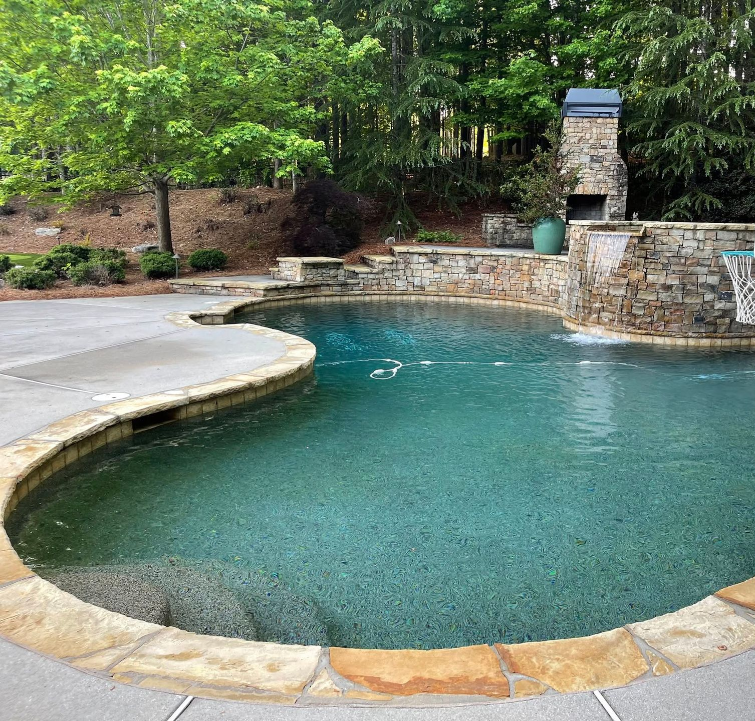 A curved backyard pool with clear blue water beside a stone wall and wooded landscaping.