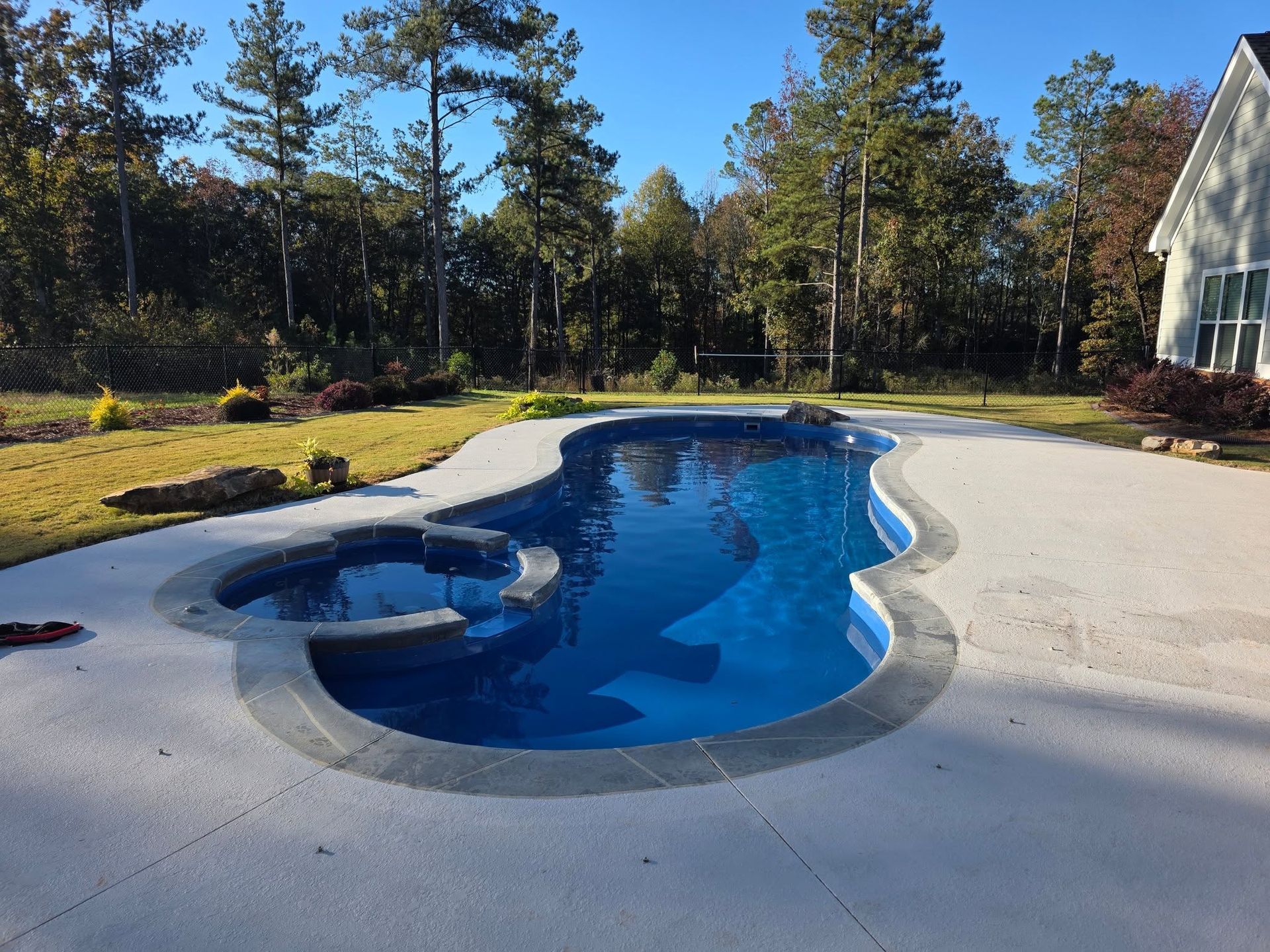 Curved backyard swimming pool with blue water, white deck, and trees under a clear sky