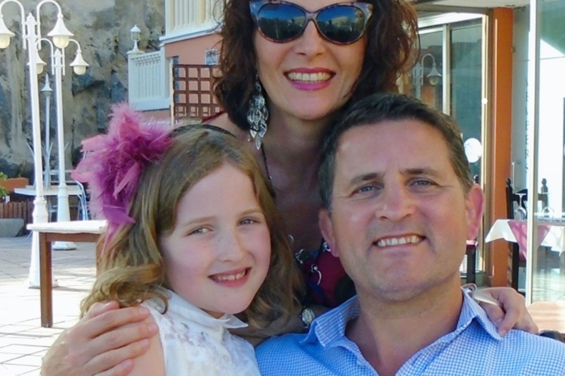 Three smiling people posing outdoors on a sunny day, with buildings and deck chairs in the background