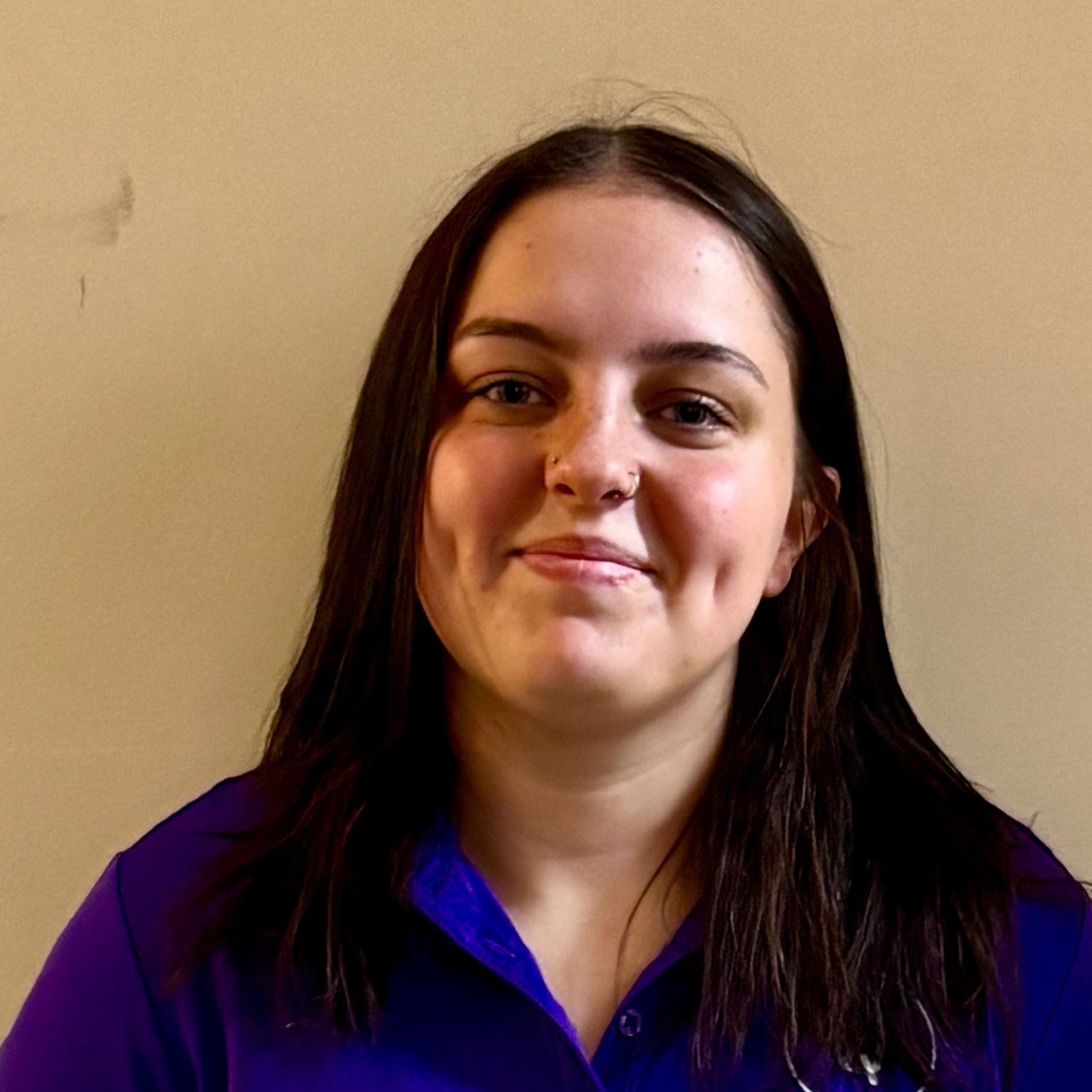 Woman with dark hair and a purple shirt, smiling against a plain beige wall