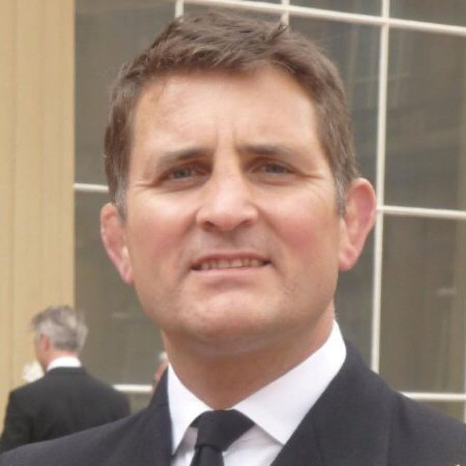 Man in a dark suit and tie standing outdoors with a neutral expression.