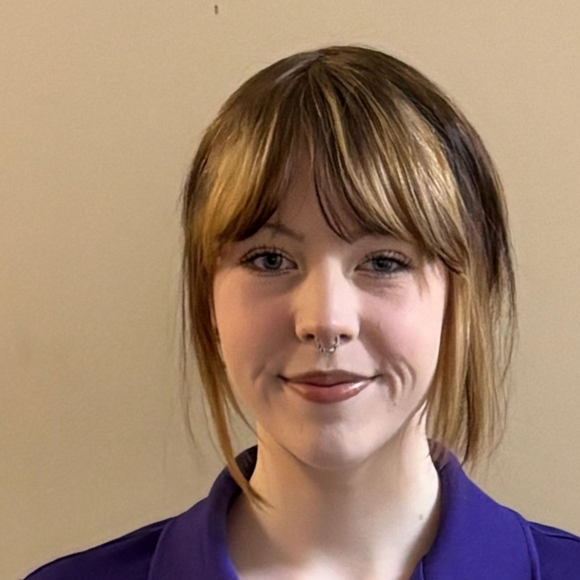 Portrait of a person with a septum ring and purple shirt against a plain beige background