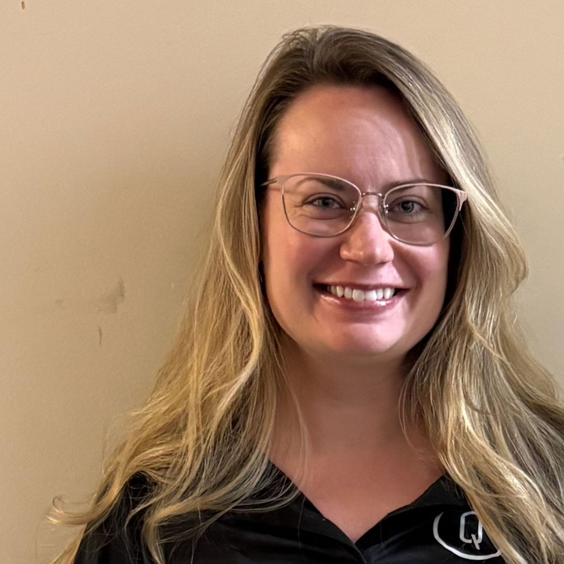 Smiling person with glasses and long blonde hair against a beige wall