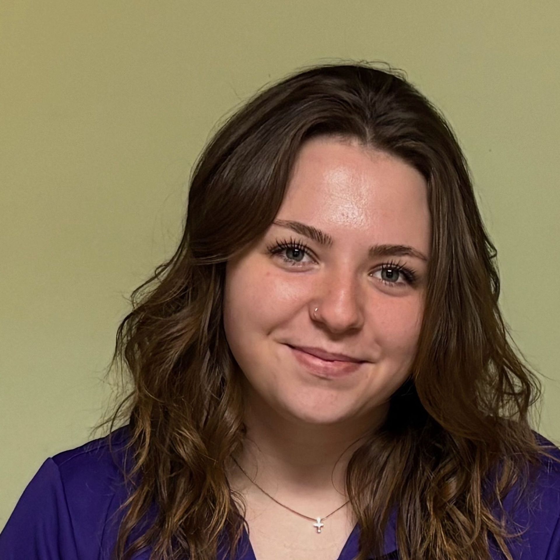 Portrait of a smiling person with wavy brown hair wearing a purple top against a plain beige background