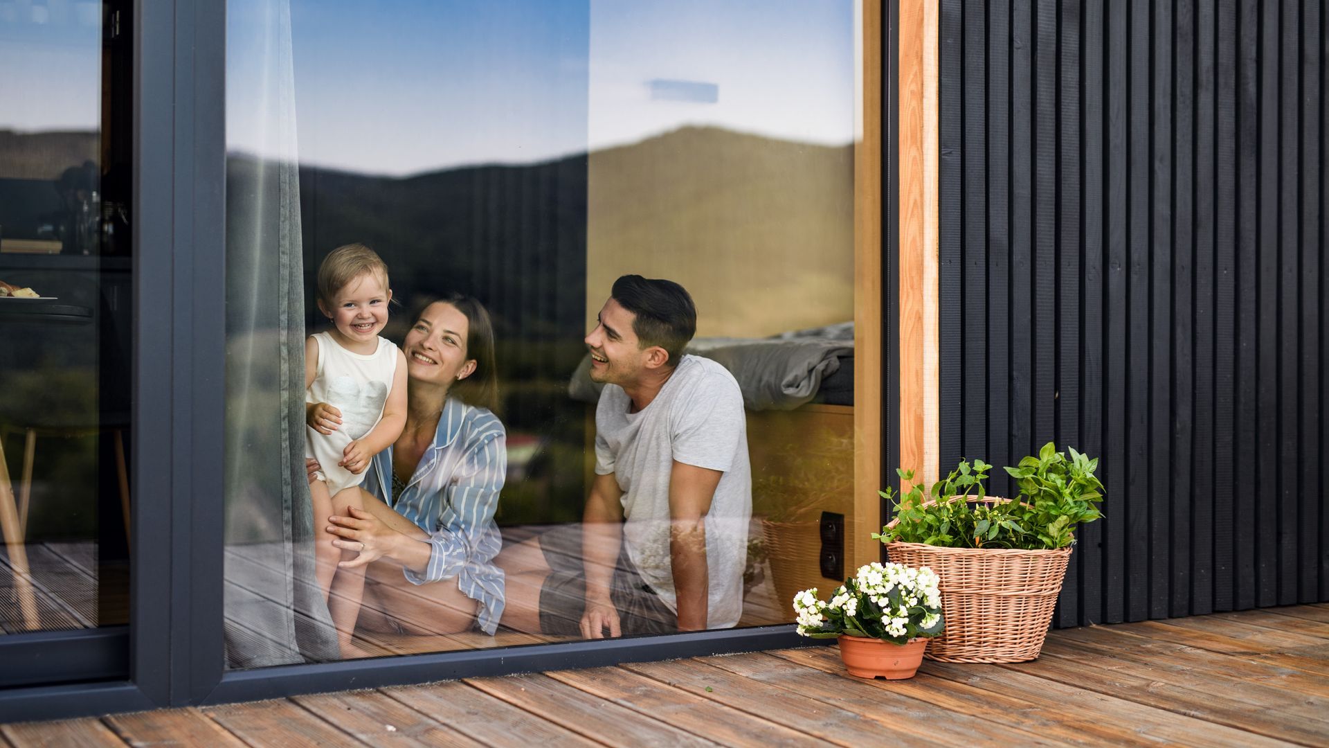 Three people sitting by a glass door, smiling inside a modern home with potted plants on the deck.