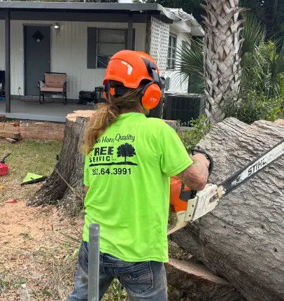 Worker in orange safety gear cutting a large tree trunk with a chainsaw outside a house.
