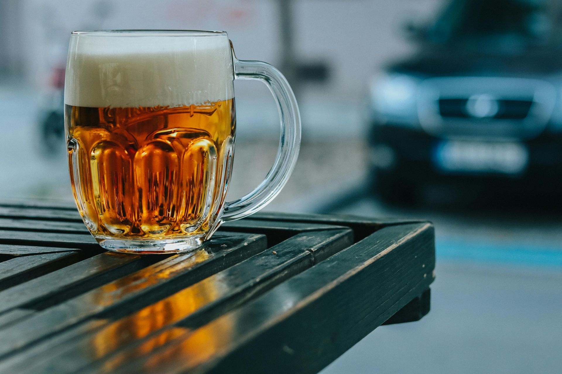 Beer mug on a wet outdoor table, with a blurred car in the background.