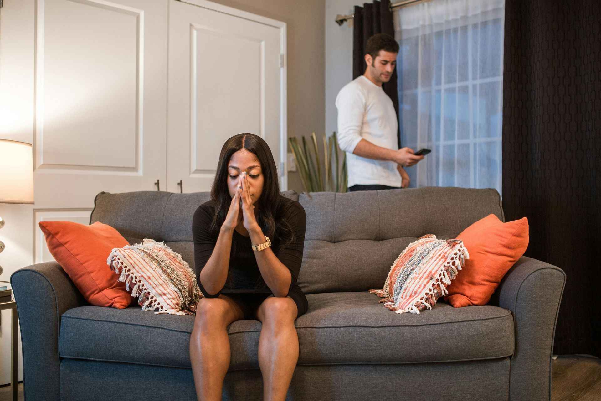 Woman sits on couch with hands clasped while man stands in background holding a plate in a living room.