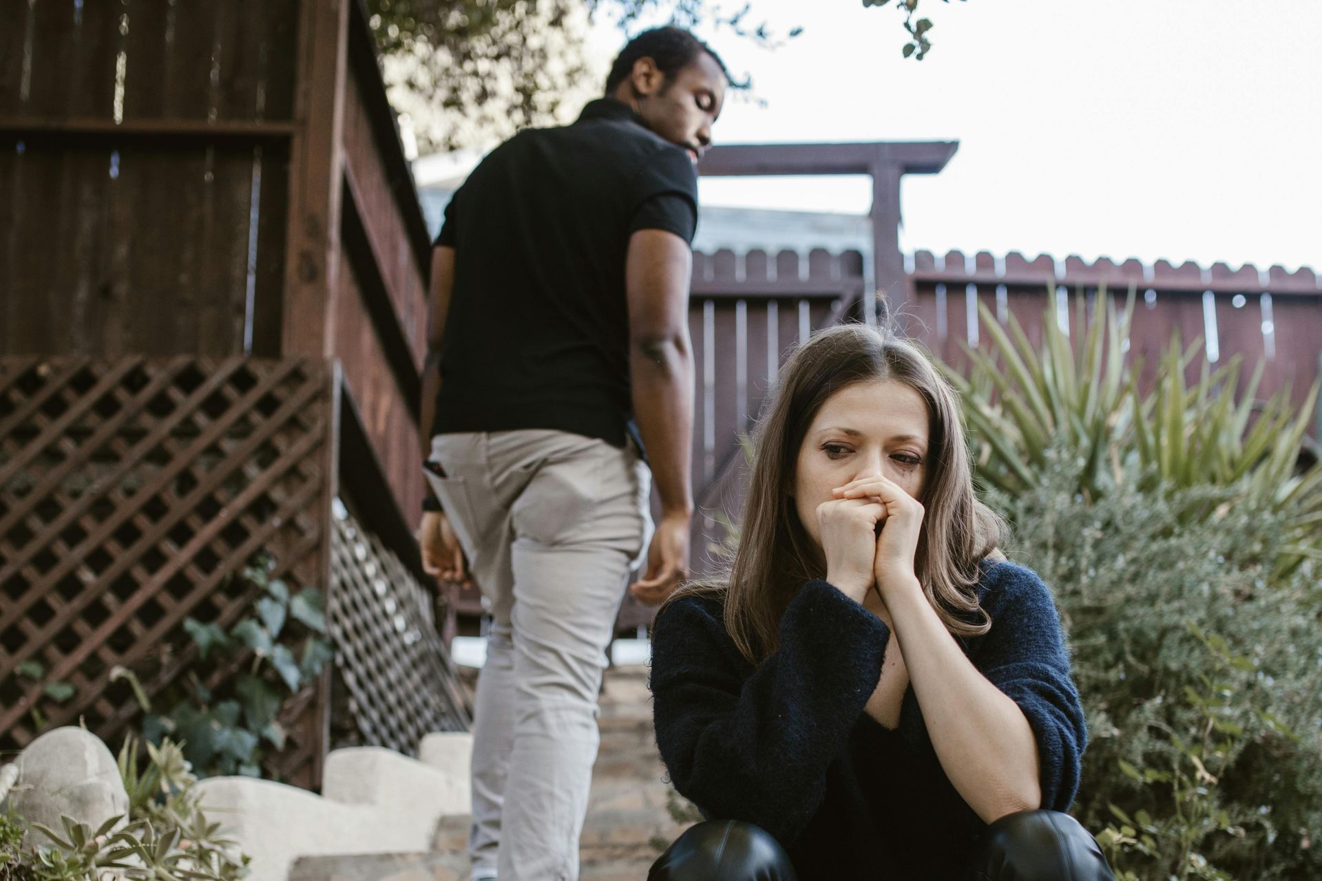A woman sits upset in a yard while a man walks away in the background.