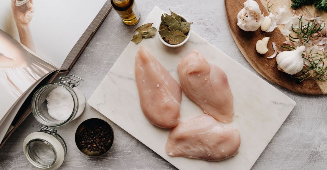 Pechugas de pollo crudas sobre una tabla de cortar, con ajo, hierbas, sal y pimienta al lado