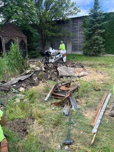 Worker using machinery to clear debris in a grassy area with a gazebo and building in the background.