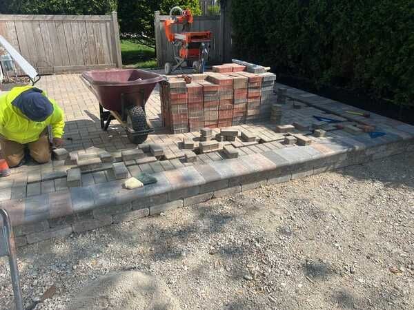 Person laying brick pavers on a patio with a wheelbarrow, bricks, and saw in the background.