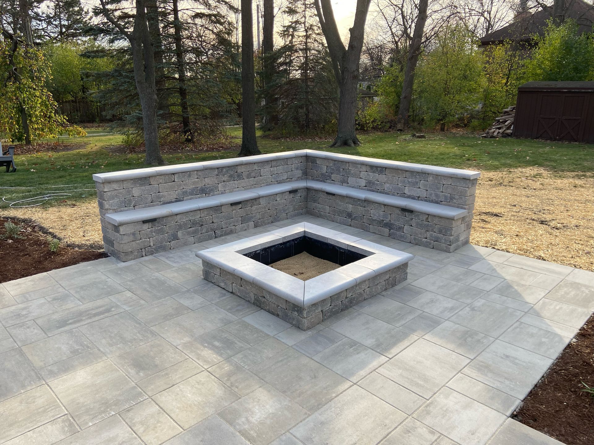 Stone fire pit with built-in bench seating on a paved patio in a yard, trees in the background.