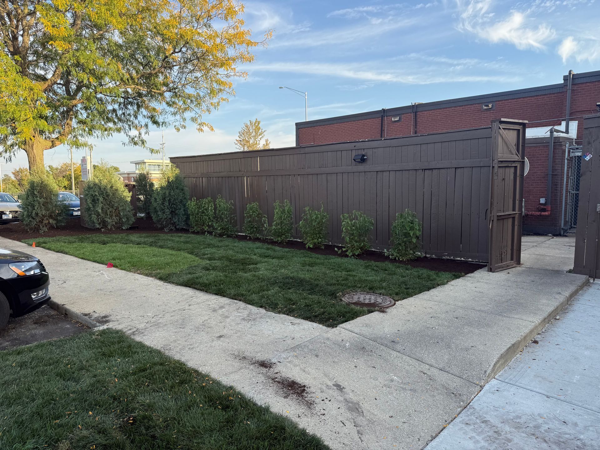 Lawn, sidewalk, and brown wooden fence outside building; shrubs line fence.