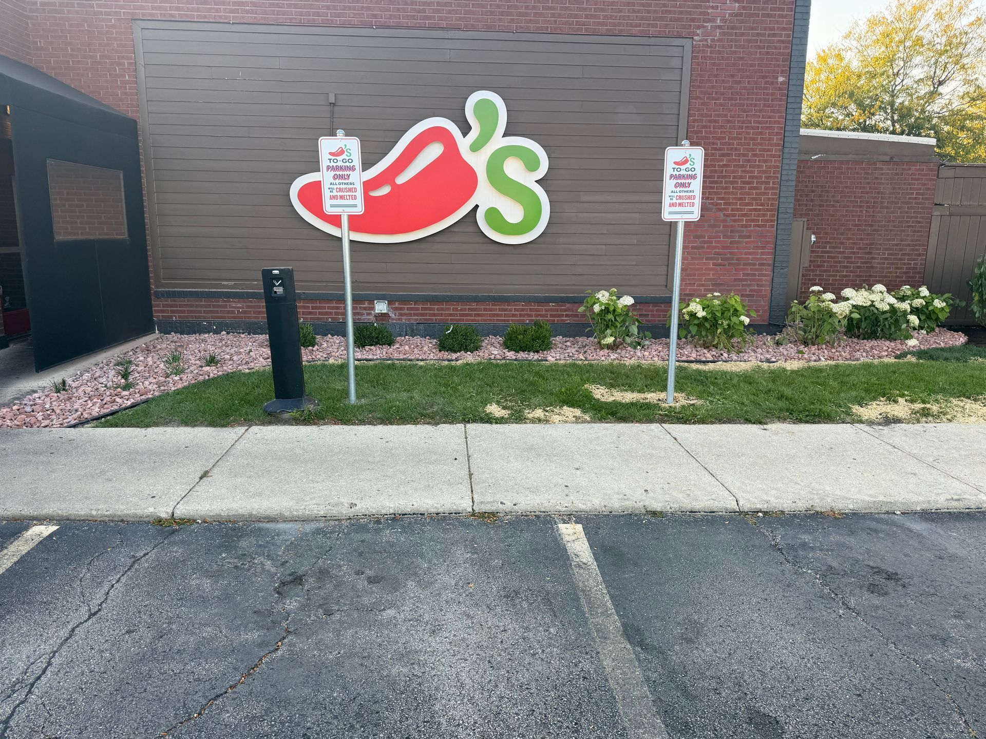 Chili's restaurant exterior with a red chili pepper logo on a brown wall, two parking signs in front, and a sidewalk.