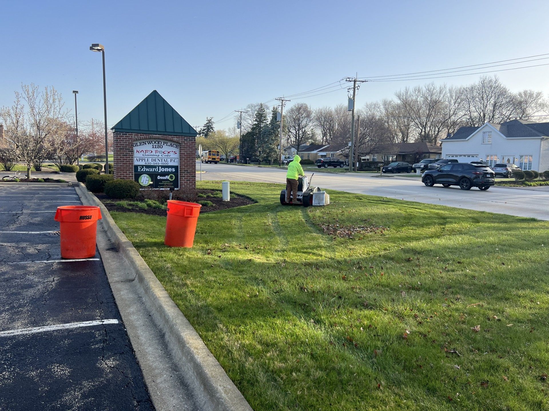 Person in yellow vest using a leaf blower on a grassy patch near a sign and a street. Orange buckets sit nearby.