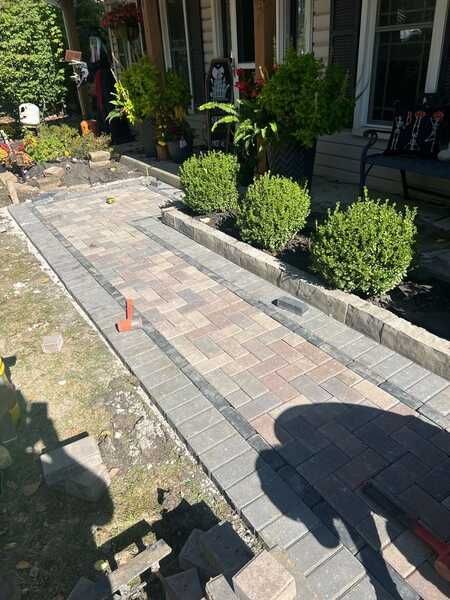 Brick pathway with borders, boxwood bushes, and potted plants in an outdoor setting.