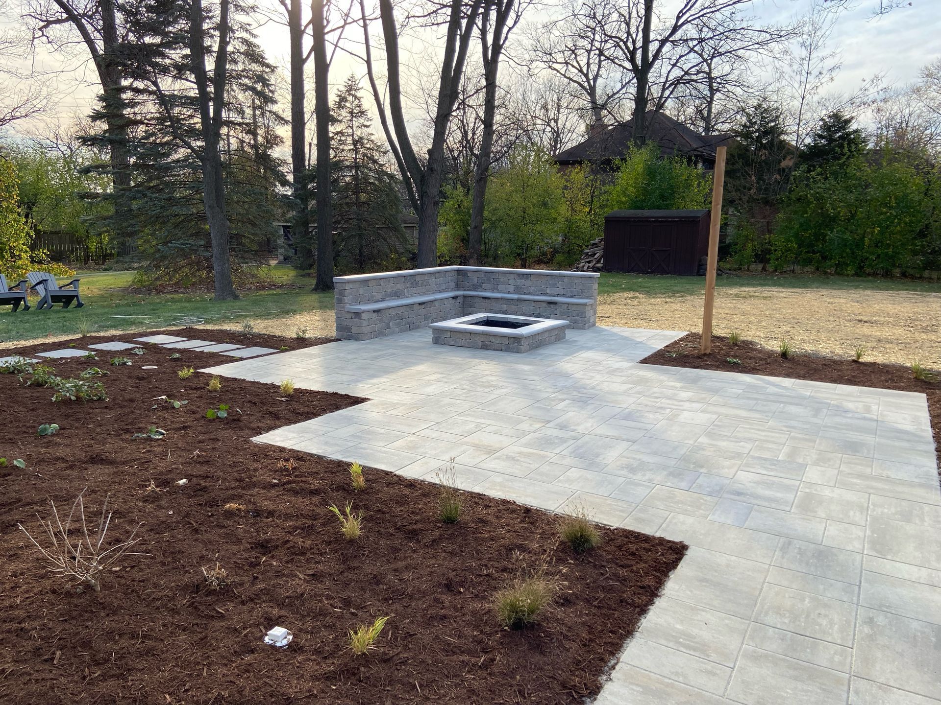 Stone patio with built-in seating around a fire pit, surrounded by mulch and trees.