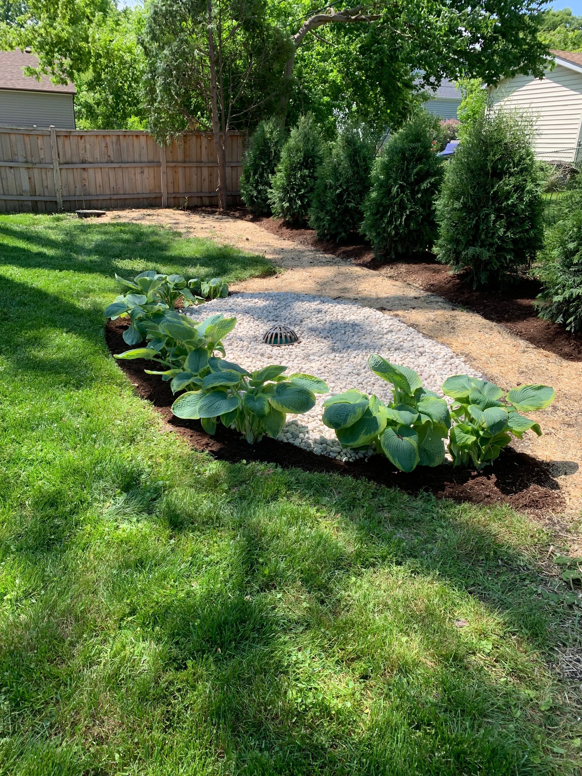 Backyard garden with green grass, brown mulch, and a gravel area surrounded by plants.
