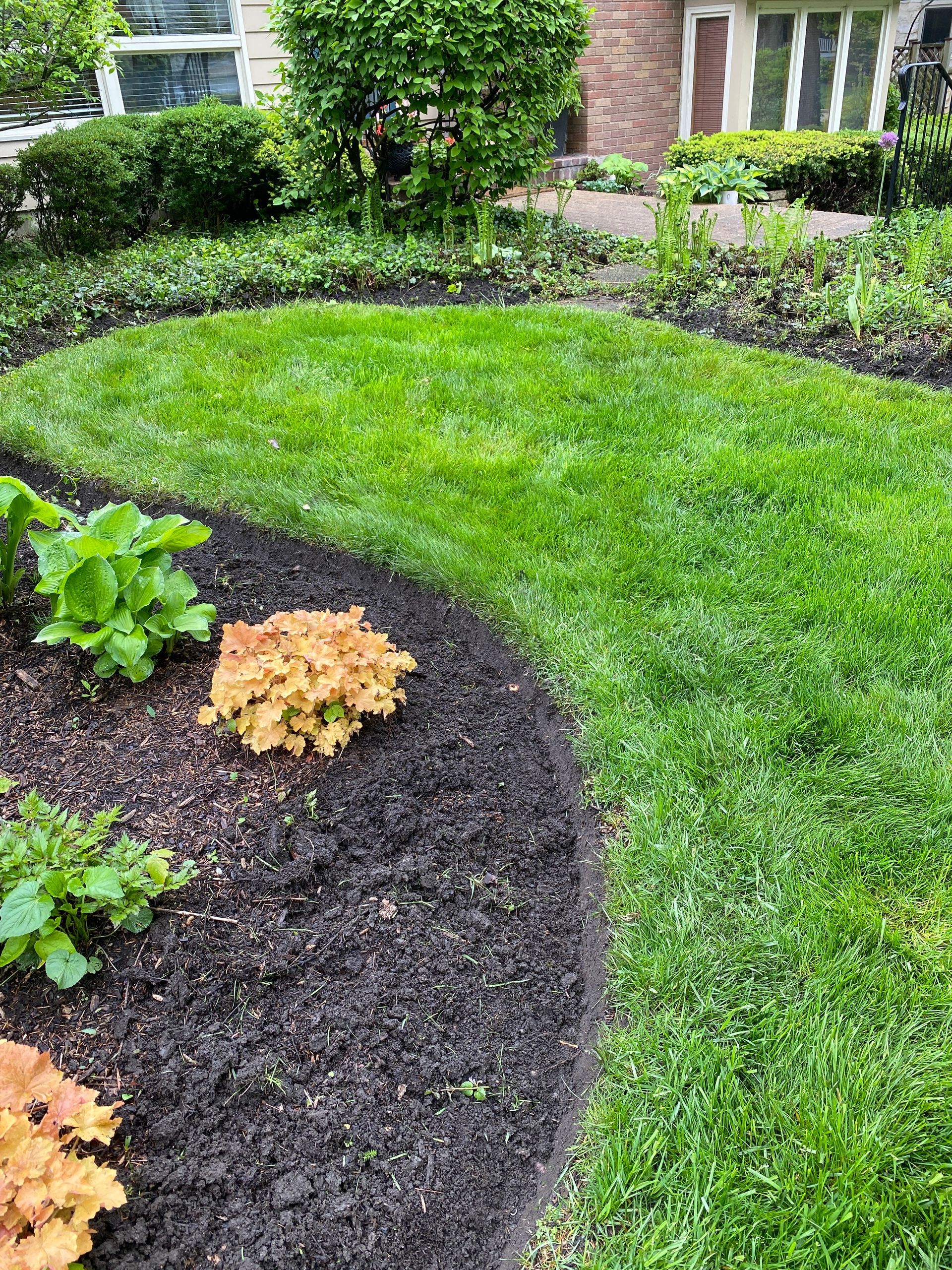 Lush green lawn borders dark mulch and colorful plants. A house with a brick facade is in the background.