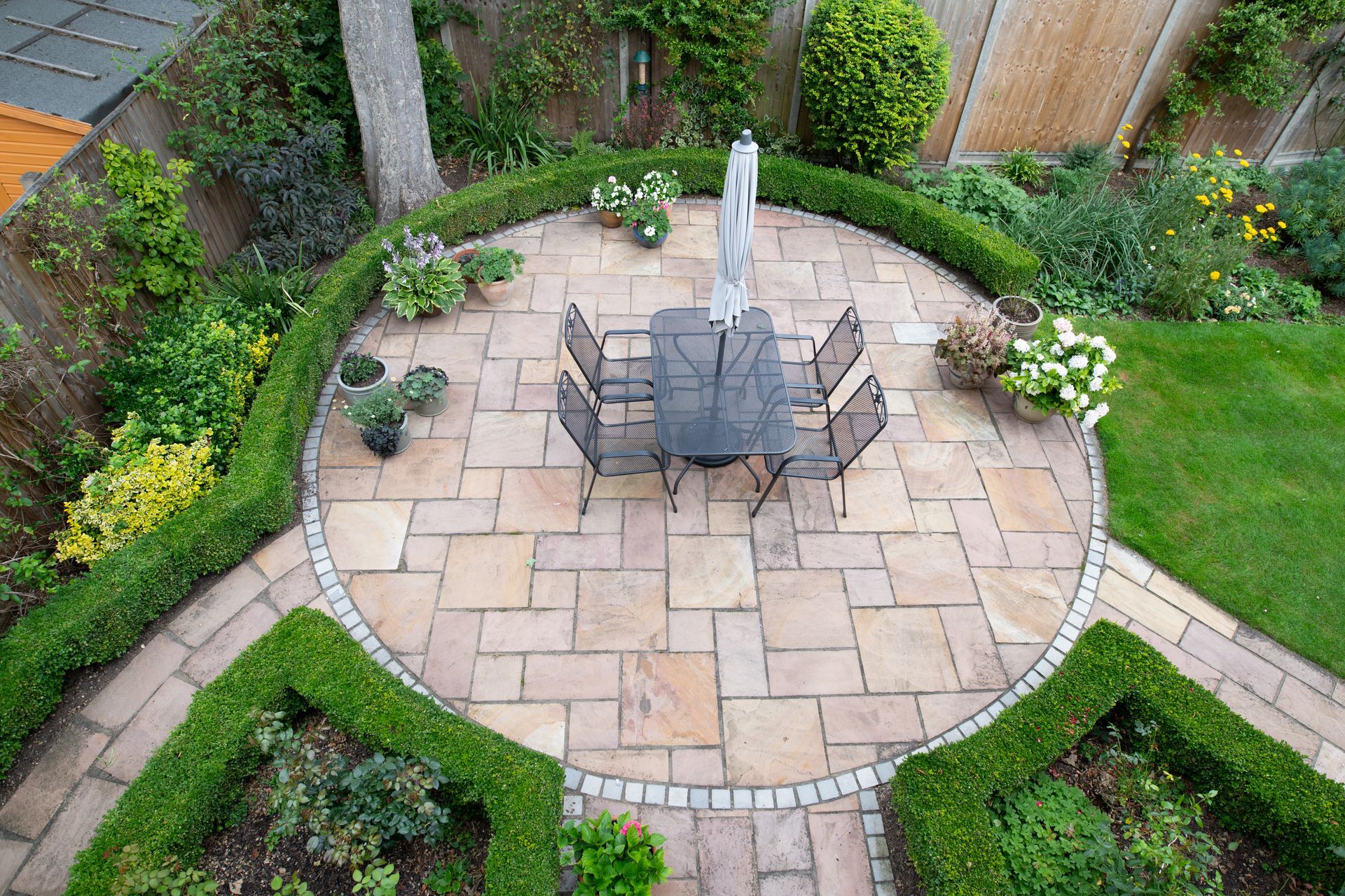 Overhead view of a circular stone patio with a table, surrounded by hedges and a garden.