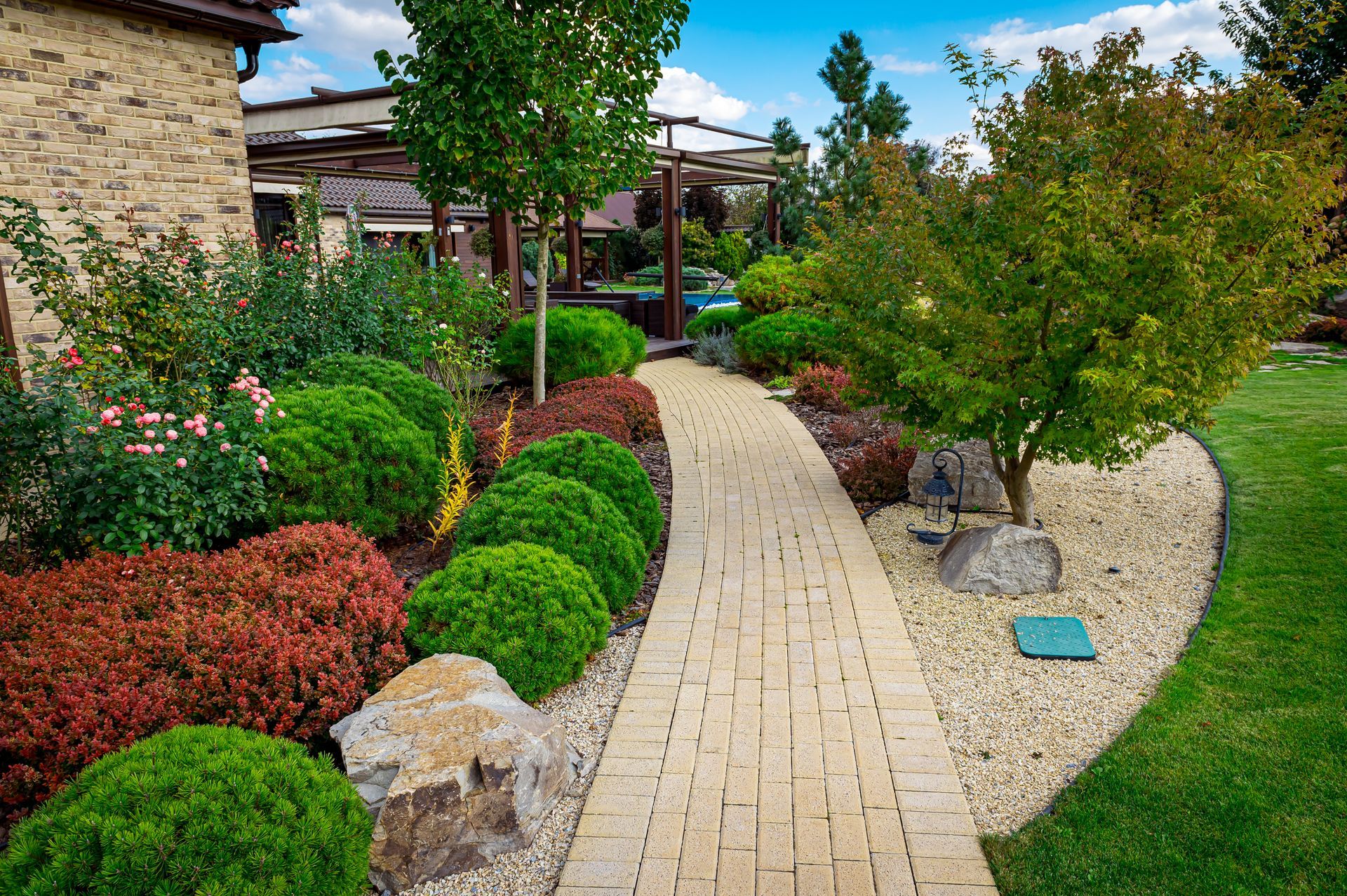 Stone pathway winds through a colorful garden with shrubs, trees, and a house in the background.