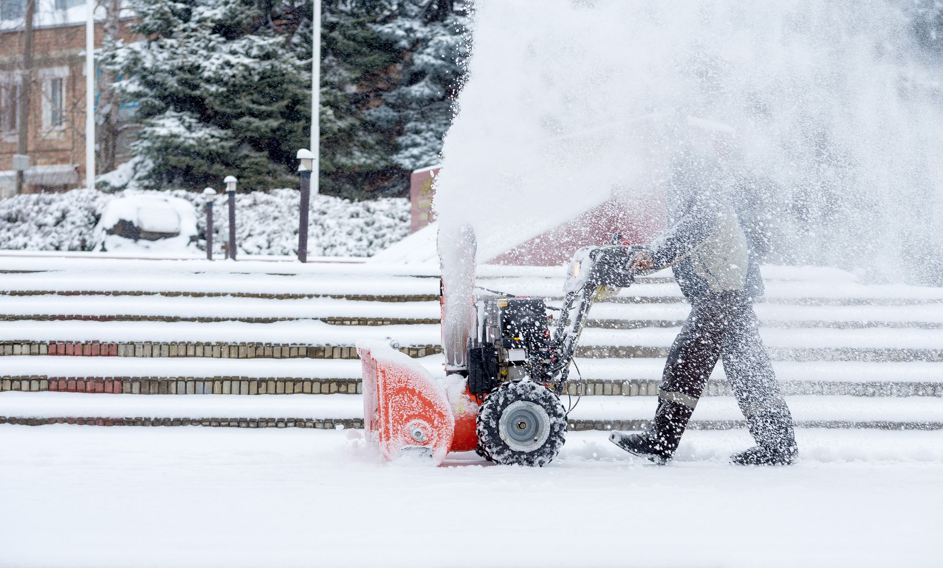 Person using an orange snowblower to clear snow from a set of outdoor steps in winter.