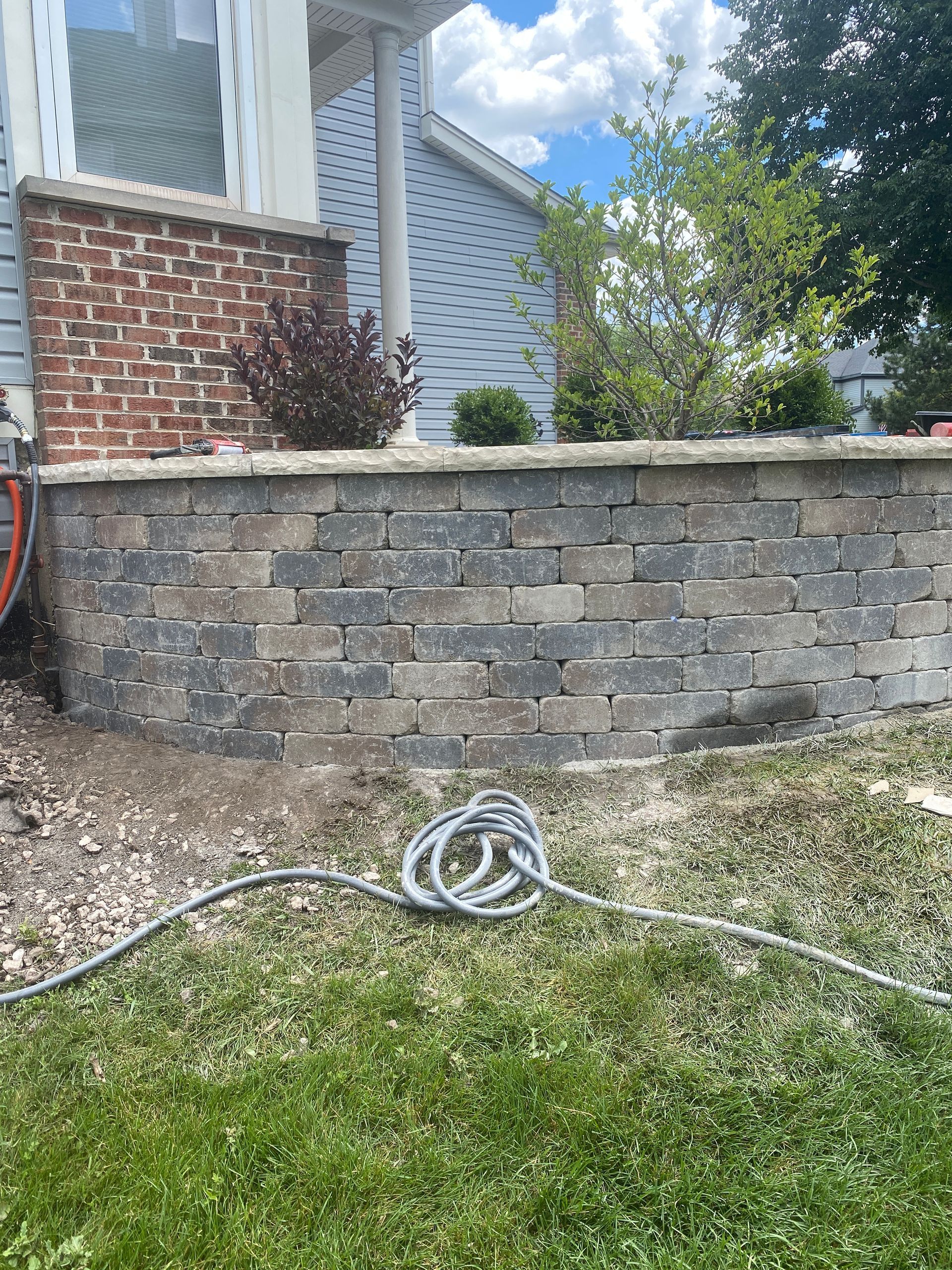 Low retaining wall made of multi-colored bricks, next to a house with blue siding. A coiled hose is on the grass.