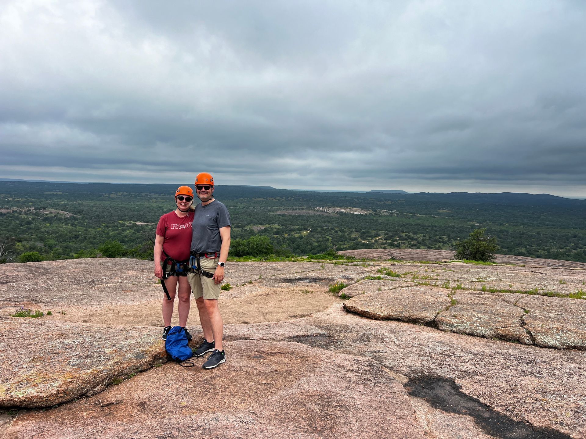 Couple wearing helmets and climbing gear pose on a mountaintop with a scenic vista under a cloudy sky.