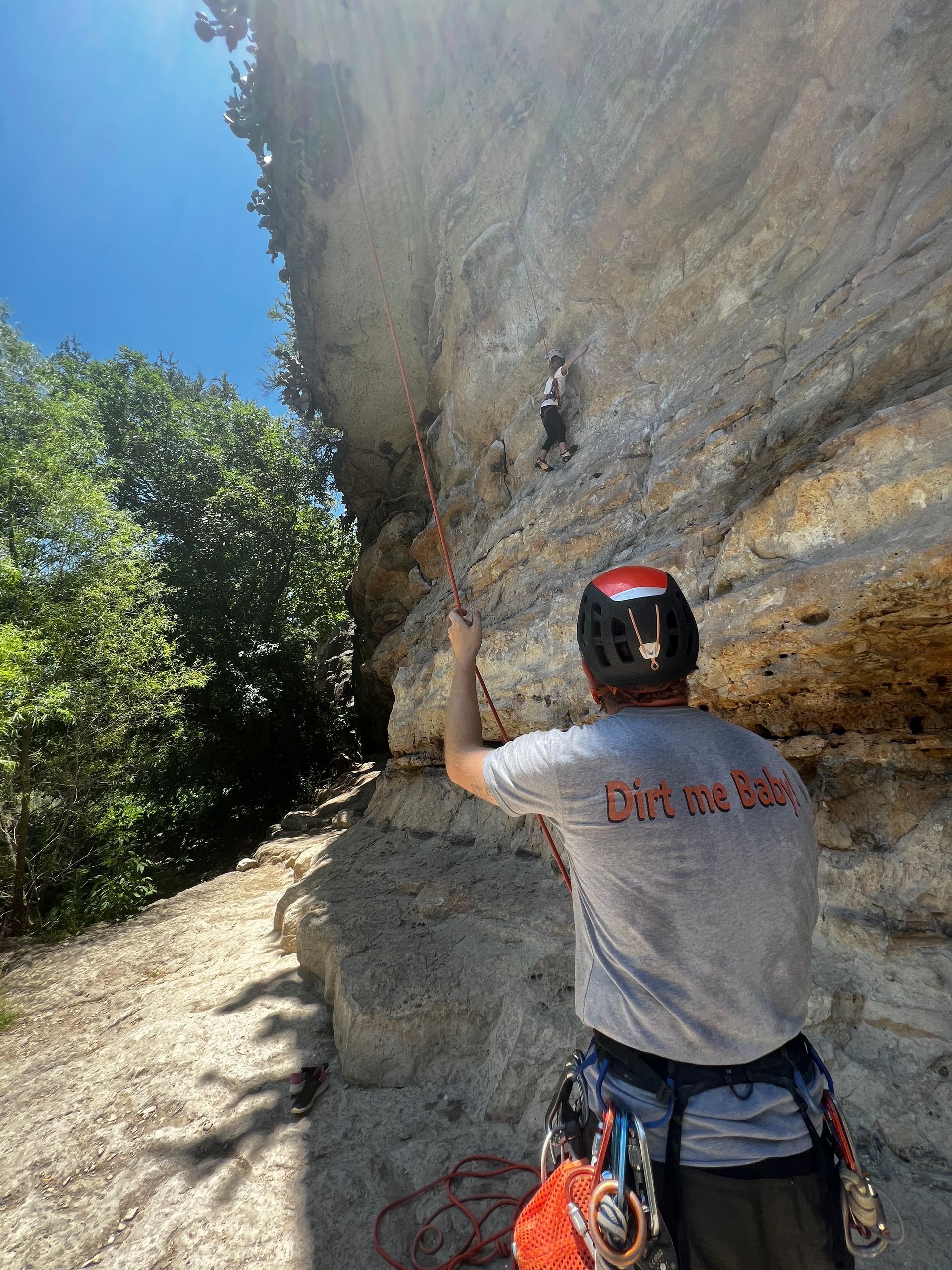 Person belaying a rock climber on a limestone cliff under a blue sky; trees in the background.