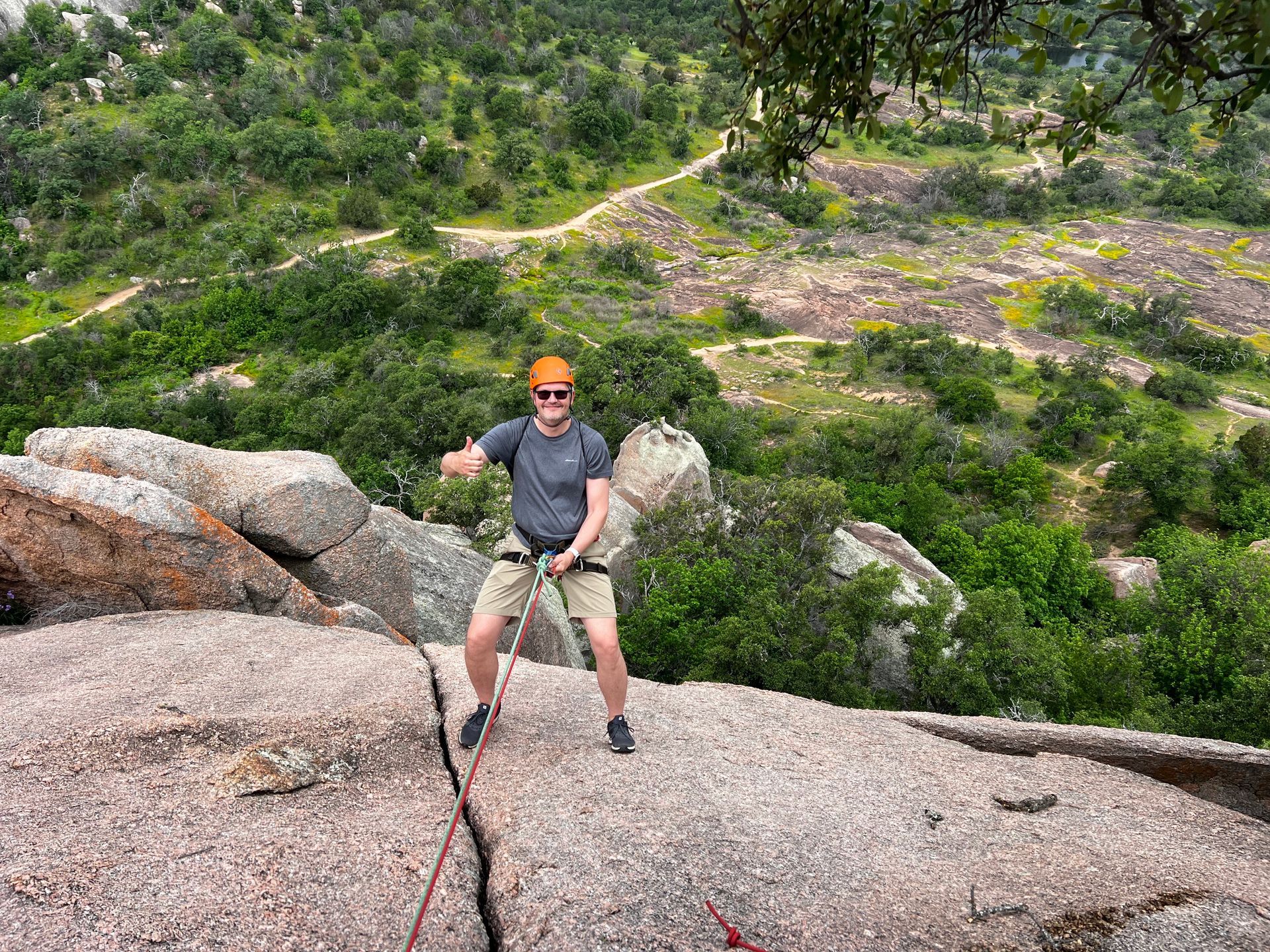 Person rappelling down a granite rock face, wearing safety gear, in a green, rocky landscape.
